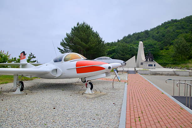 Gangneung, South Korea - June 1st 2025: A retired training jet with red markings sits on display near a military monument surrounded by greenery at Gangneung Unification Park.