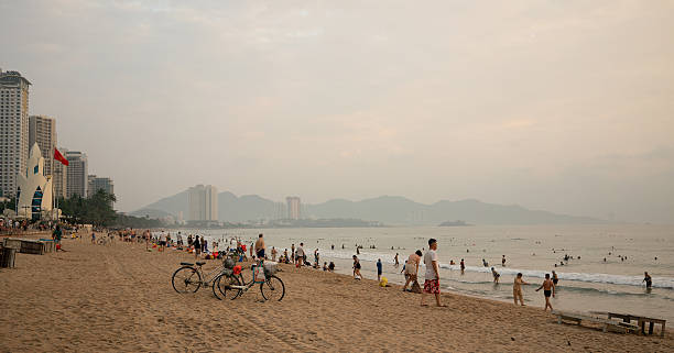 Nha Trang, Vietnam - April 08, 2025: a panoramic view of Nha Trang beach at sunrise, with tire tracks, bicycles, swimmers, and distant city buildings.