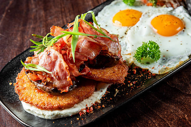 istock Delicious breakfast with sunny-side-up eggs, crispy hash browns, grilled bacon, and fresh herbs, served on a dark plate with rustic wooden 2223068560