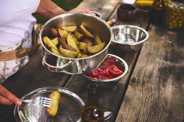 A person prepares and serves roasted potato wedges with fresh tomato slices on a rustic outdoor table, evoking a picnic-like setting.