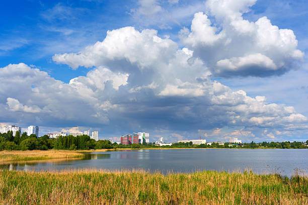 Clear view of the Kyiv skyline with buildings reflecting in a tranquil lake, surrounded by green vegetation and dramatic clouds overhead.