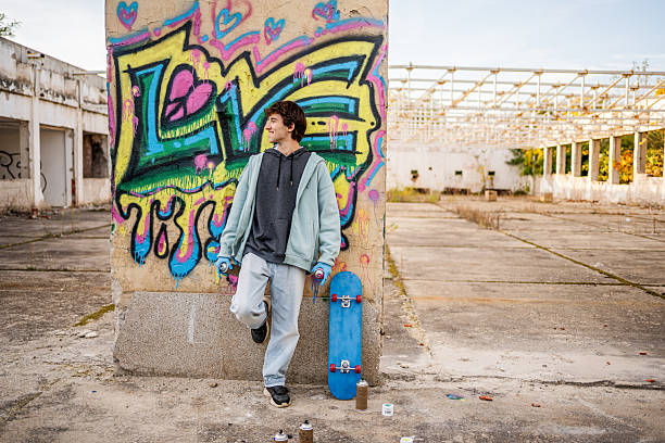 Young man with spray cans and a skateboard stands beside a colorful graffiti wall in an abandoned urban setting
