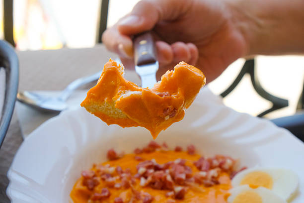 a man, sitting at a table, is about to eat a piece of bread, held on a fork, dipped in spanish salmorejo, also known as porra antequerana, a cold tomato soup, topped with boiled egg and chopped jamon