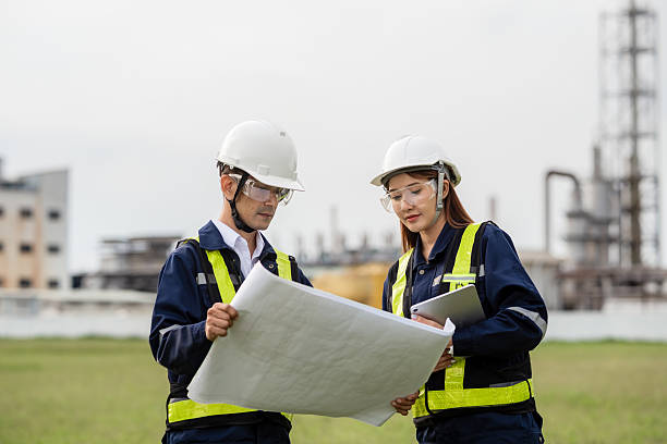 istock Engineering Team Analyzing Blueprint and Tablet Data at an Industrial Site. Two people wearing safety gear and looking at a drawing 2223093285