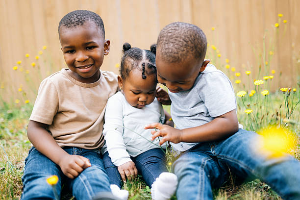 African American children sit together in a yard, picking and examining flowers.  Cute brother and sister interactions.