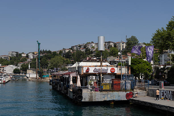 ISTANBUL, TURKEY - JULY 3: A small, aged ferry dock covered with signage, graffiti, and utility structures is seen on the Bosphorus shoreline on July 3, 2025, in Istanbul, Turkey. The dock appears worn and heavily used, reflecting the functional nature of Istanbul’s local maritime infrastructure. In the background, residential buildings spread across a hillside, while purple flags add visual interest to the waterfront scene. The setting captures the layered textures of urban life in Istanbul’s coastal neighborhoods.