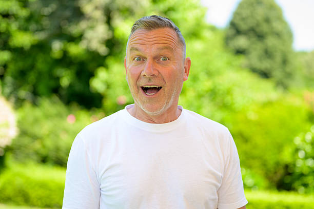 Cheerful senior man in a white t-shirt expressing surprise with wide open eyes and mouth, standing outdoors in a green garden.