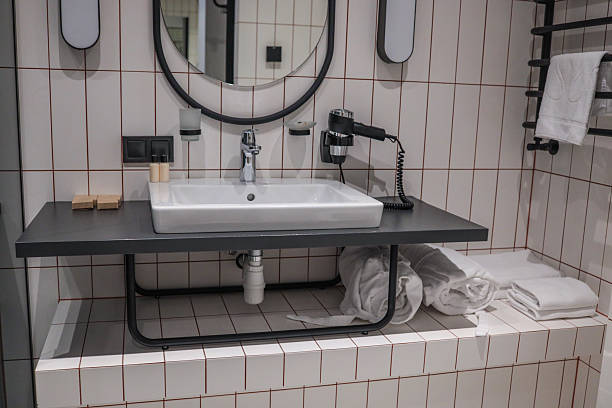 istock Modern hotel bathroom interior featuring oval mirror, white-tiled walls, sleek countertop basin, chrome faucet, wall-mounted hairdryer and neatly rolled towels for a spa-like ambience 2223100911