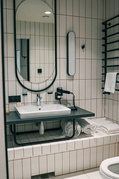 istock Modern hotel bathroom interior featuring oval mirror, white-tiled walls, sleek countertop basin, chrome faucet, wall-mounted hairdryer and neatly rolled towels for a spa-like ambience 2223100919