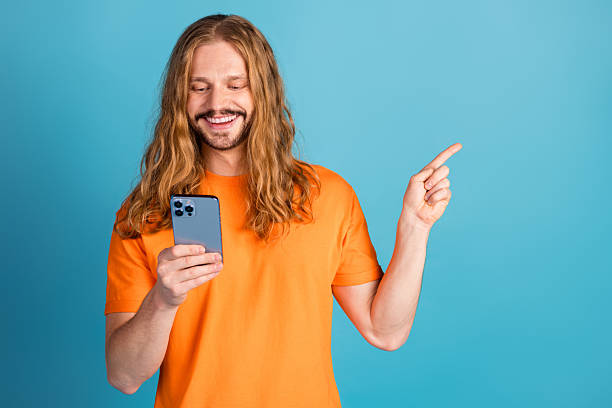 istock Young man with long hair smiling at smartphone while gesturing against blue background in casual bright orange shirt 2223105647