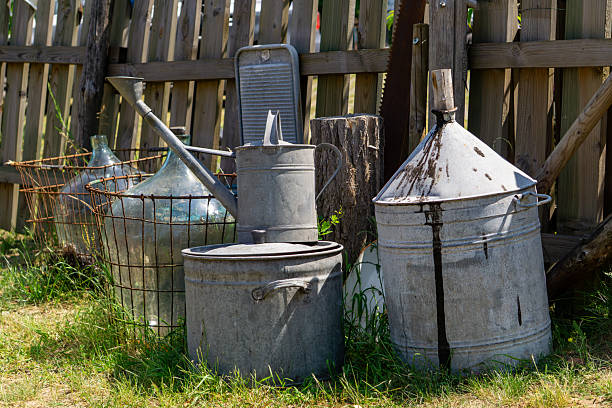 vintage galvanized garden tools and containers placed by a wooden fence. Includes a watering can, washboard, barrel, demijohns in wire baskets, and other rustic items. Countryside setting with warm summer sunlight.