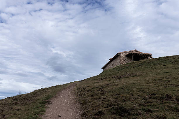 Winding hiking trail ascends a grassy hill towards a solitary stone hut nestled on the hilltop, beneath a cloudy sky