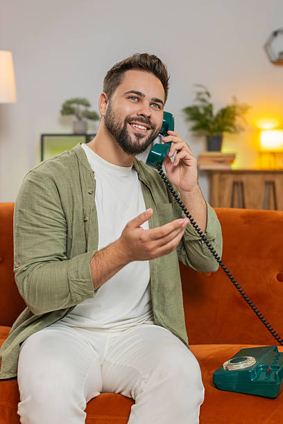 istock Smiling young man making wired telephone call conversation with friends sitting on sofa at home 2223118659