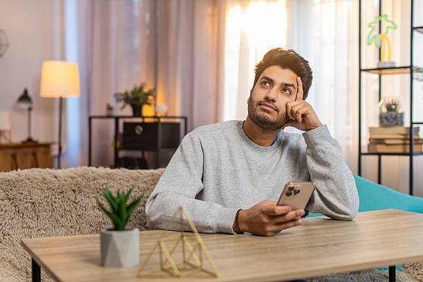 istock Young Indian man texting on smartphone while resting head on hand at table in living room at home 2223121821