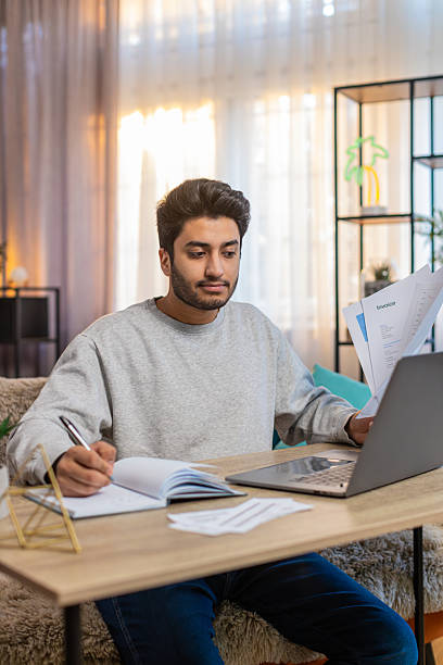 istock Young Indian man freelancer reviewing financial reports on laptop while taking notes in home office 2223121825