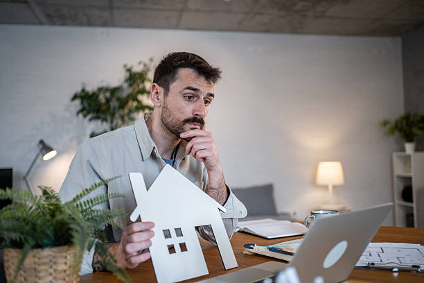 istock Architect holding a small house model while thinking at home office desk 2223122569