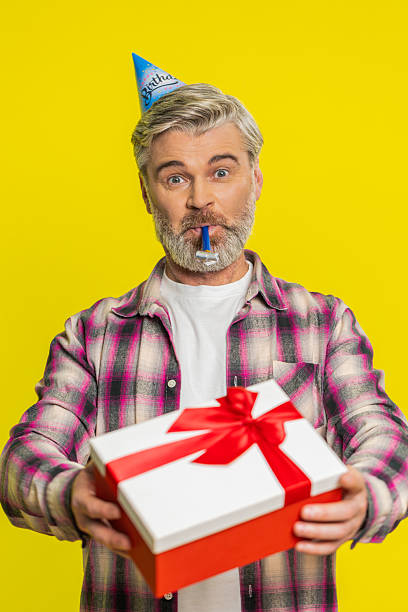 istock Mature man handing presenting birthday gift box cone hat blower joyful expression festive atmosphere 2223127512