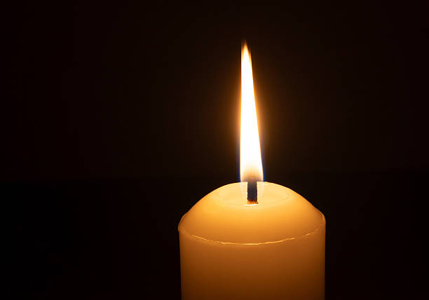istock A single burning candle flame or light glowing on a big white candle isolated on black or dark background on table in church for Christmas, funeral or memorial service 2223154467