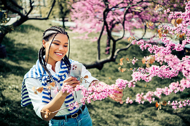 istock Young woman enjoying springtime outdoors, smiling while capturing blooming cherry blossoms on camera in a city park 2223176900