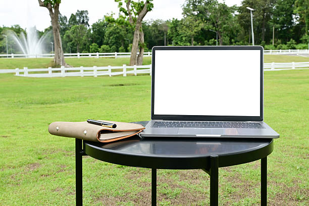A laptop is open on a table in a grassy field. The laptop is empty and the table is surrounded by grass. the laptop and notebook as the only objects on the table