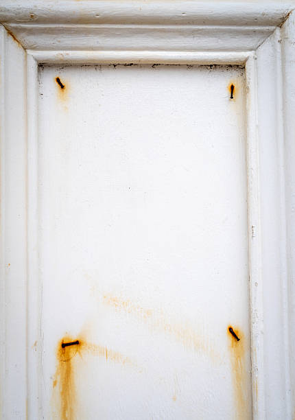 A close-up of a white wooden frame with rusty nails and peeling paint. The surface is aged, weathered, and marked by corrosion and natural wear