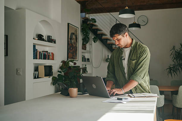 istock Young man using a laptop while standing and working from kitchen counter at home with natural light. 2223189282