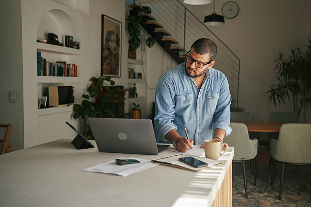 istock Man looking at laptop while writing as he  works from home standing at a counter in a modern living space 2223189678