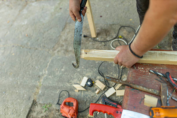 Senior man using electric planer on wooden plank outdoors, surrounded by various hand tools. DIY home improvement and woodworking concept. Close up photo