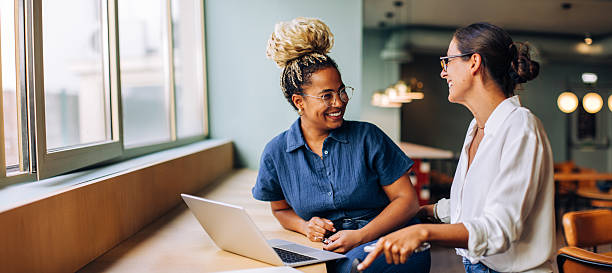 Two young women smiling and talking while working together at a laptop Two women are seated in a bright indoor setting, engaging in conversation near a laptop. They appear to be sharing ideas and enjoying their discussion while wearing casual attire. women leader stock pictures, royalty-free photos & images