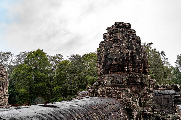 A giant stone face tower from the Bayon Temple stands prominently under a rainy sky, surrounded by tropical forest. The ancient Khmer architecture, representing Avalokiteshvara or King Jayavarman VII, reflects the grandeur of the Angkor era. The moss-covered surface and wet stones enhance the mystical atmosphere of this UNESCO World Heritage Site.