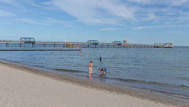 Young boy sitting barefoot on a wooden pier. Dressed in a striped t-shirt and green shorts, he smiles at the camera, the vast turquoise-colored sea stretching behind him. \nShot with Nikon D800