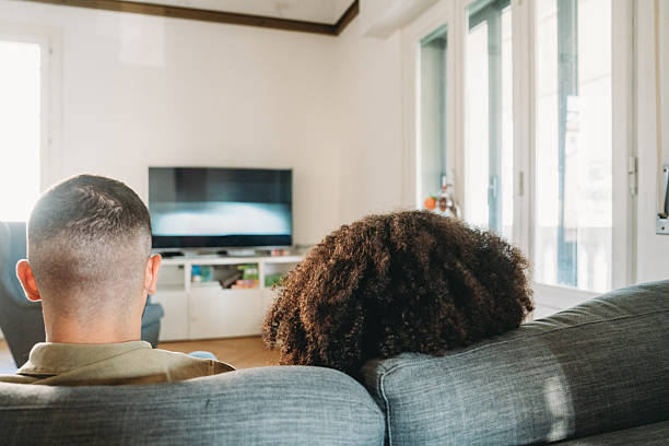 Back view of an interracial couple relaxing on a sofa, enjoying a television show together in a cozy living room filled with natural light