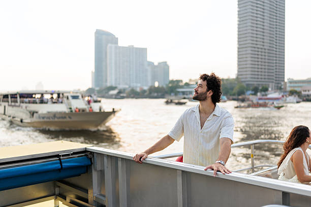 A handsome man stands on a boat, and looking up at the sky. Other boats pass by on the Chao Phraya river, with Bangkok’s tall buildings in the background.