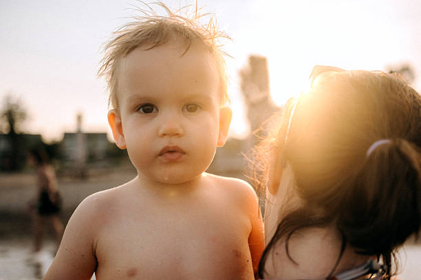 Adorable young boy smiling at the camera as his mother holds him near the water’s edge. Warm and loving family moment at the beach