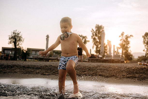 Young boy wearing snorkeling goggles, standing still in clear, shallow sea water. Calm and curious moment during beach playtime.