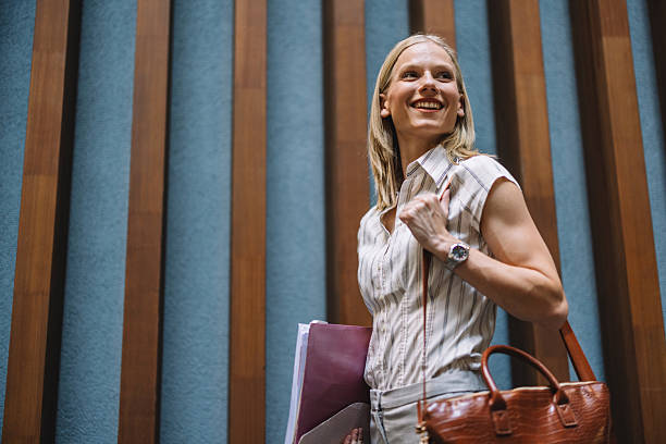 A confident woman with a smile, holding files and a handbag, represents professionalism and achievement in a modern office setting amidst stylish wooden decor.