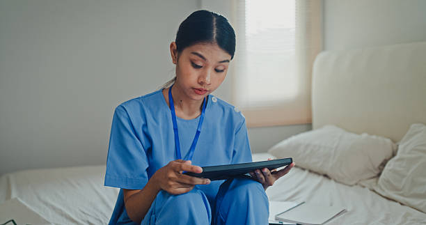 istock Young Asian female nurse in blue scrubs sitting on a bed while using a digital tablet. Nurse lifestyle concept. 2223232531