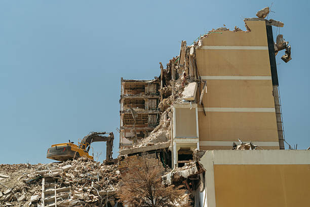 Demolition site with a collapsed building, exposed rebar, and a bulldozer working amid concrete rubble and dust. Apartment building destroyed by an earthquake or accident.