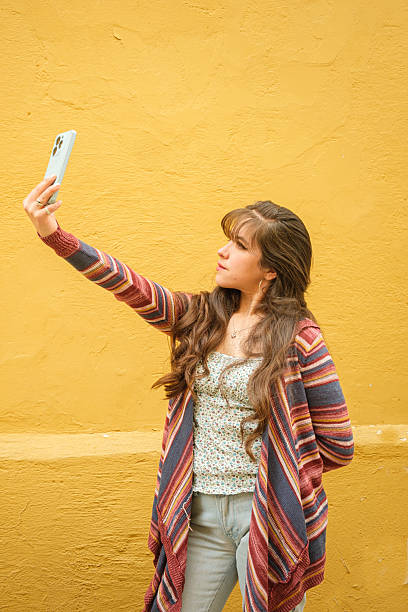 istock vertical white-skinned latina woman taking a selfie with her cell phone on a wall with a yellow background - modeling concept 2223237016
