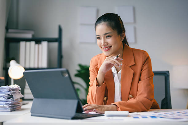 istock Efficient young asian businesswoman sitting at her desk, happily using a digital tablet with paperwork and a calculator nearby, indicating a productive workspace 2223249593