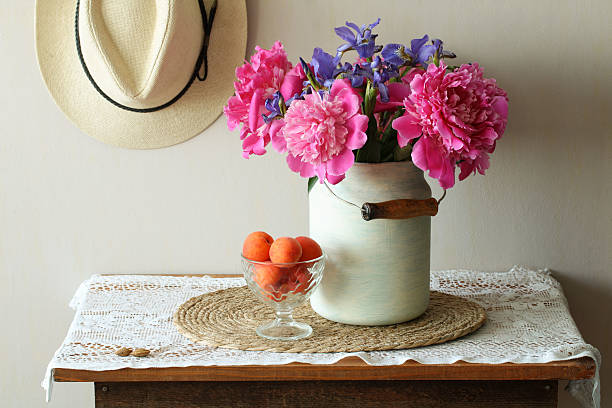 Bouquet of pink peonies and irises in vintage milk can, glass bowl of apricots, straw placemat and doily on rustic wooden table with hat on wall