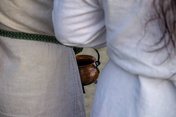 figures in medieval-style clothing, featuring linen tunics and a copper cauldron hanging from a belt. Historical reenactment scene focused on costume details.