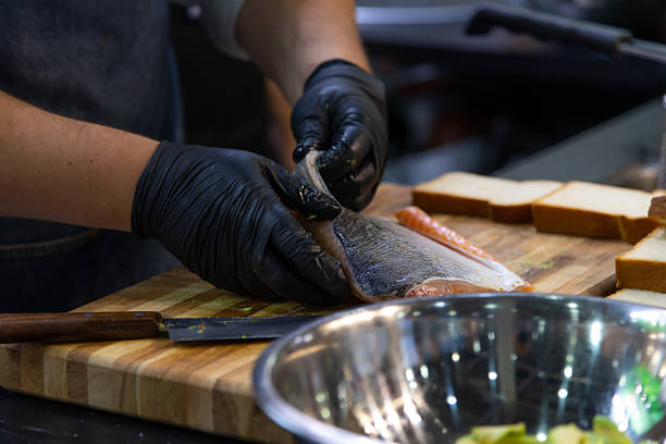 A cook skillfully fillets fish while wearing gloves in a busy kitchen. Nearby, sliced bread is ready to complement the dish, showcasing a vibrant culinary preparation scene.