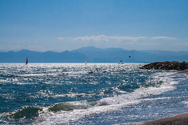 Picturesque view of sea waves sparkling in the sun with mountains on the horizon. Sailboats and parasailers are seen on the water capturing a peaceful coastal moment in summer.