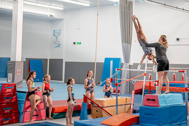 A coach guides a young gymnast performing an advanced uneven bars routine as other students watch, showcasing teamwork and skill development in a gymnasium setting.