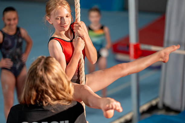 A young gymnast works on strengthening skills during a rope climbing exercise, guided by a coach. The training scene emphasizes determination, athleticism, and teamwork in a gymnasium environment.