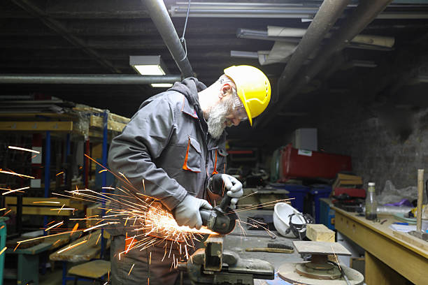 Senior Industrial Worker With A Beard Grinding Metal In A Workshop While Wearing A Yellow Hard Hat, Safety Goggles, And Gloves. Bright Sparks Fly As He Uses An Angle Grinder On A Steel Pipe. Concept: Heavy Industry, Skilled Labor, Safety, Experience.