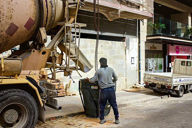 Sliema, Malta - 04/15/2025: A construction worker operates a cement mixer truck on a city street, filling a container with concrete. Urban construction site with trucks, tools, and buildings in the background.