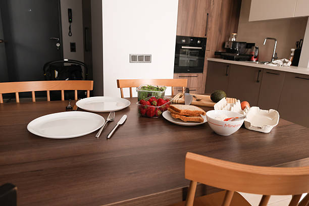 Sliced avocado, greens, bread, and herbal tea are being prepared and served on a wooden kitchen table with plates and cutlery.
