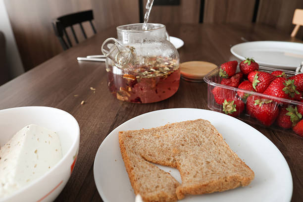 Sliced avocado, greens, bread, and herbal tea are being prepared and served on a wooden kitchen table with plates and cutlery.